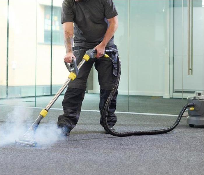A man steam cleaning a grey carpet in the middle of  an empty office with glass walls