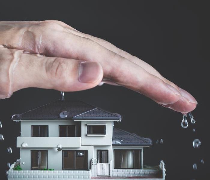 A wet hand hovering over a small model home as water continues to pour on top of the hand and small model home